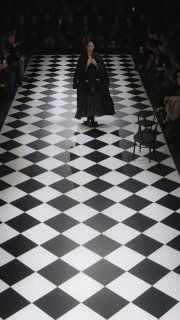 Панорамный кадр подиума: Ирина Шейк в чёрном платье на показе Junya Watanabe, шахматный пол и публика по краям зала