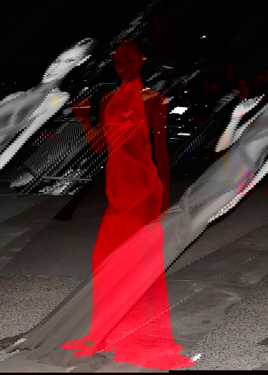 Белла Хадид покидает место премьеры в красном Schiaparelli: струящееся платье, вечерняя улица и поклонники вдалеке создают контекст события.