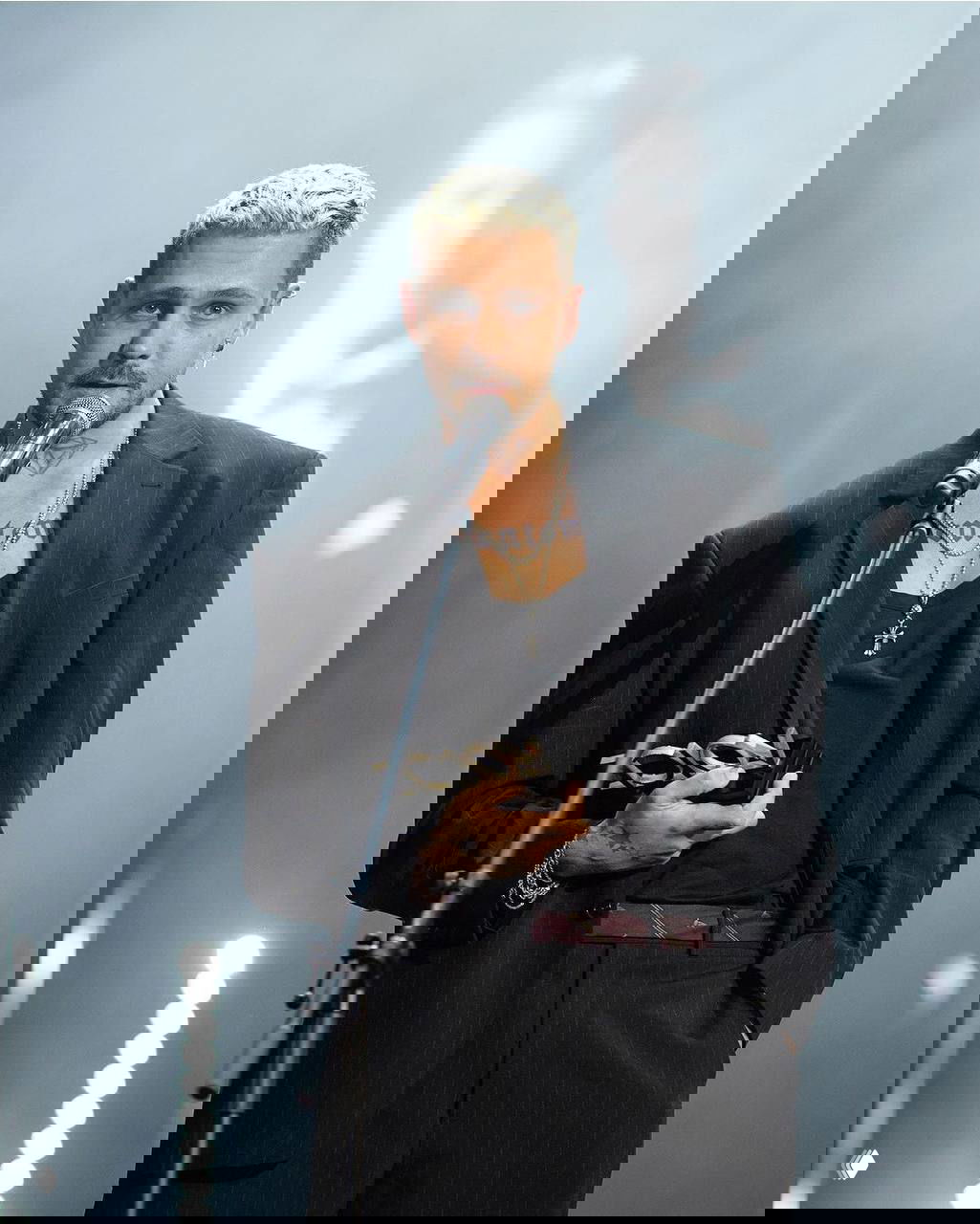 Милош Бикович на сцене с микрофоном, сценическое освещение, татуировки и серьёзный образ во время публичного выступления.