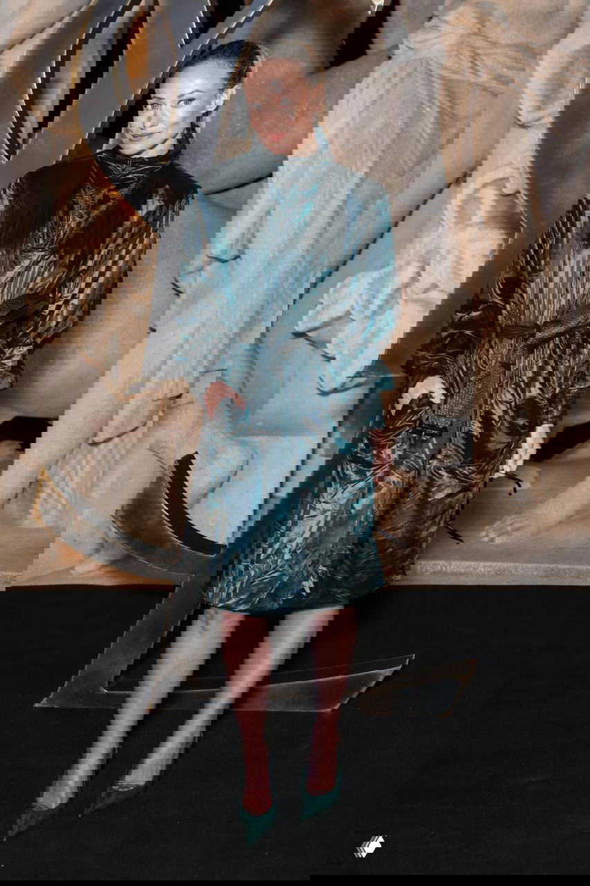 Гостья в тёмно-зелёном блестящем плаще с поясом и туфлях позирует на чёрном ковре на фоне барельефной скульптуры и логотипа YSL.