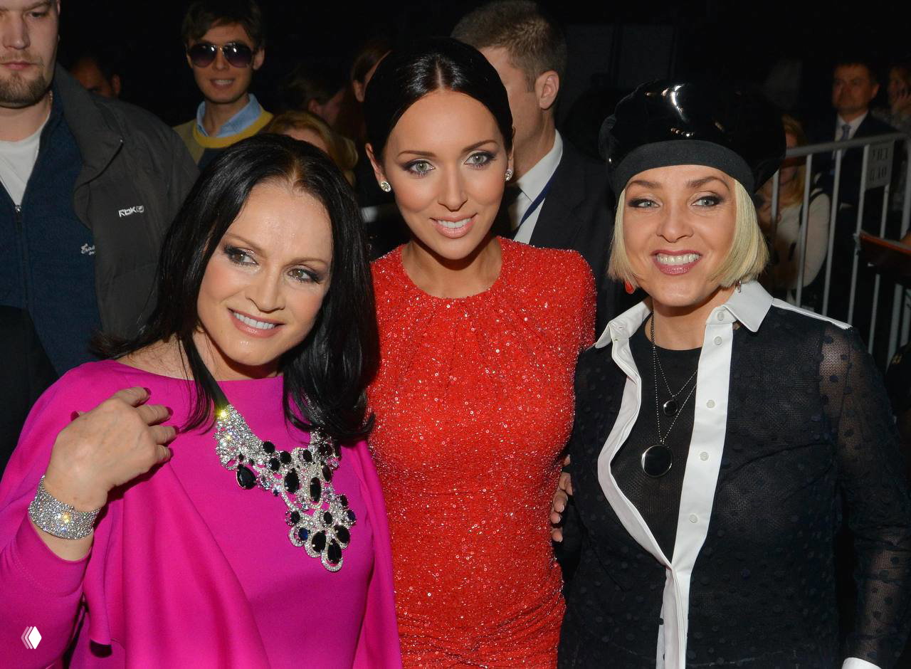 Три знаменитости, включая Лайму Вайкуле, на светском приёме: яркие наряды, вечерняя сцена и камеры вокруг, дружеская и праздничная атмосфера.