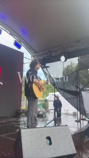 Антон Токарев на сцене сап‑фестиваля «ЭТО САП, Москва!»: поёт с акустической гитарой под навесом; видны микрофоны, колонка, звуковики и дождливая обстановка.