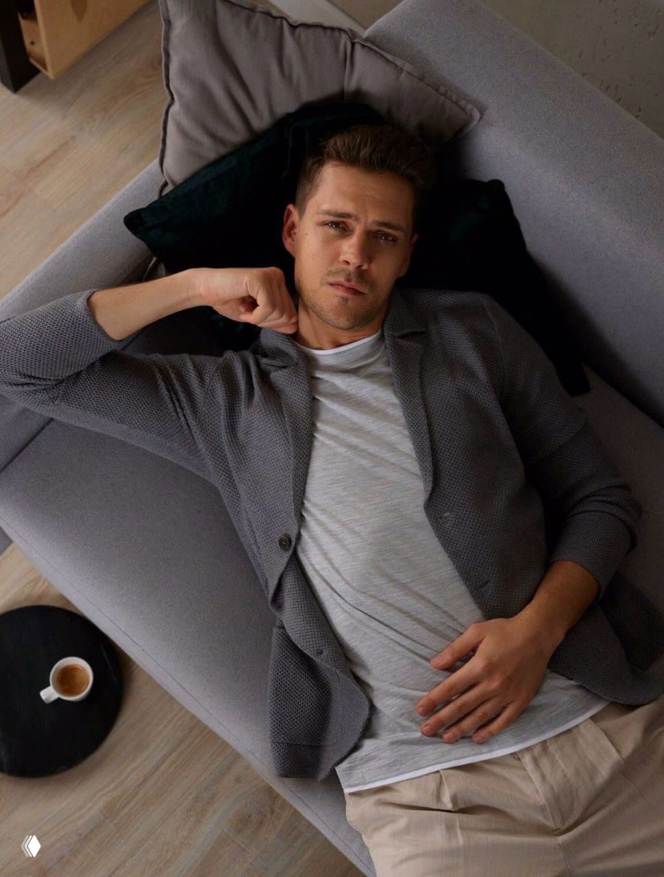 Милош Бикович отдыхает на диване, расслабленная поза в домашней обстановке, кадр в интерьерном стиле с чашкой на столике.