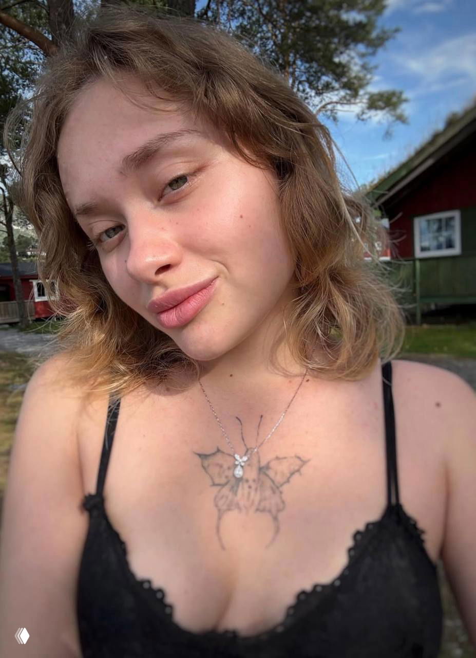 Крупный план лица и верхней части туловища Марии Ковальчук на улице возле домика в Норвегии: естественный вид, татуировка бабочки на груди хорошо видна.