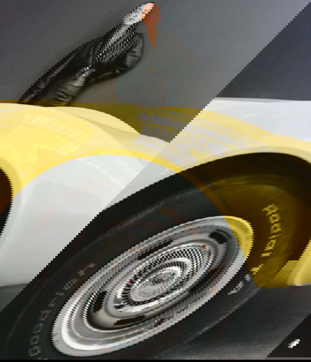 Фотография модели позади крупного жёлтого спортивного автомобиля: акцент на колесе, контраст цветов и ретро‑атмосфера съёмки.