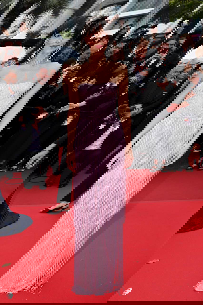 Женщина в пастельно-розовом платье в стиле бахромы, длина в пол, открытые плечи; образ запечатлён на красной дорожке с фотографами на заднем плане.