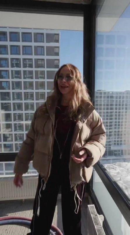 Самбурская у панорамного окна квартиры с видом на соседний фасад; видно светлую лоджию и городской пейзаж за стеклом.