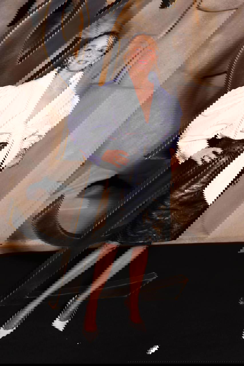 Женский образ с объёмной белой блузой и чёрной кожаной юбкой, широкий ремень и белые лодочки на фоне логотипа Saint Laurent во время мероприятия.