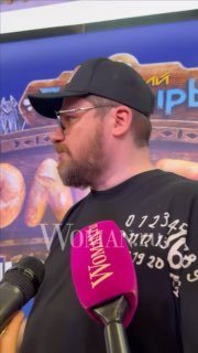 Профиль Гарика Харламова в кепке и бородой, рядом микрофон Woman.ru, он смотрит в сторону, фон студии размыт.