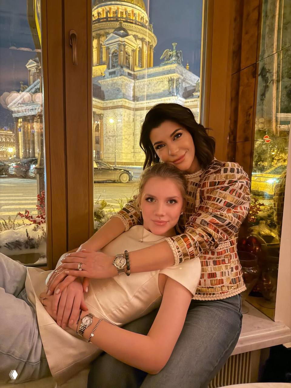 Екатерина Волкова обнимает подругу, сидящую на подоконнике; тёплые тона и ночной городской пейзаж видны за окном, кадр передаёт камерную атмосферу вечера.