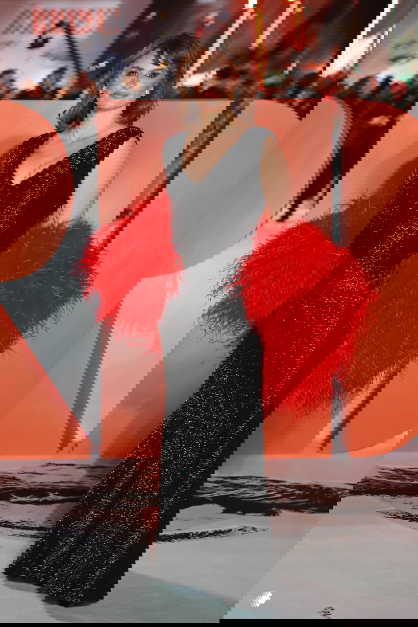 Полный рост женщины в черном платье и объемных красных перьевых аксессуарах на фоне оранжевых декораций и гостей премьеры фильма.