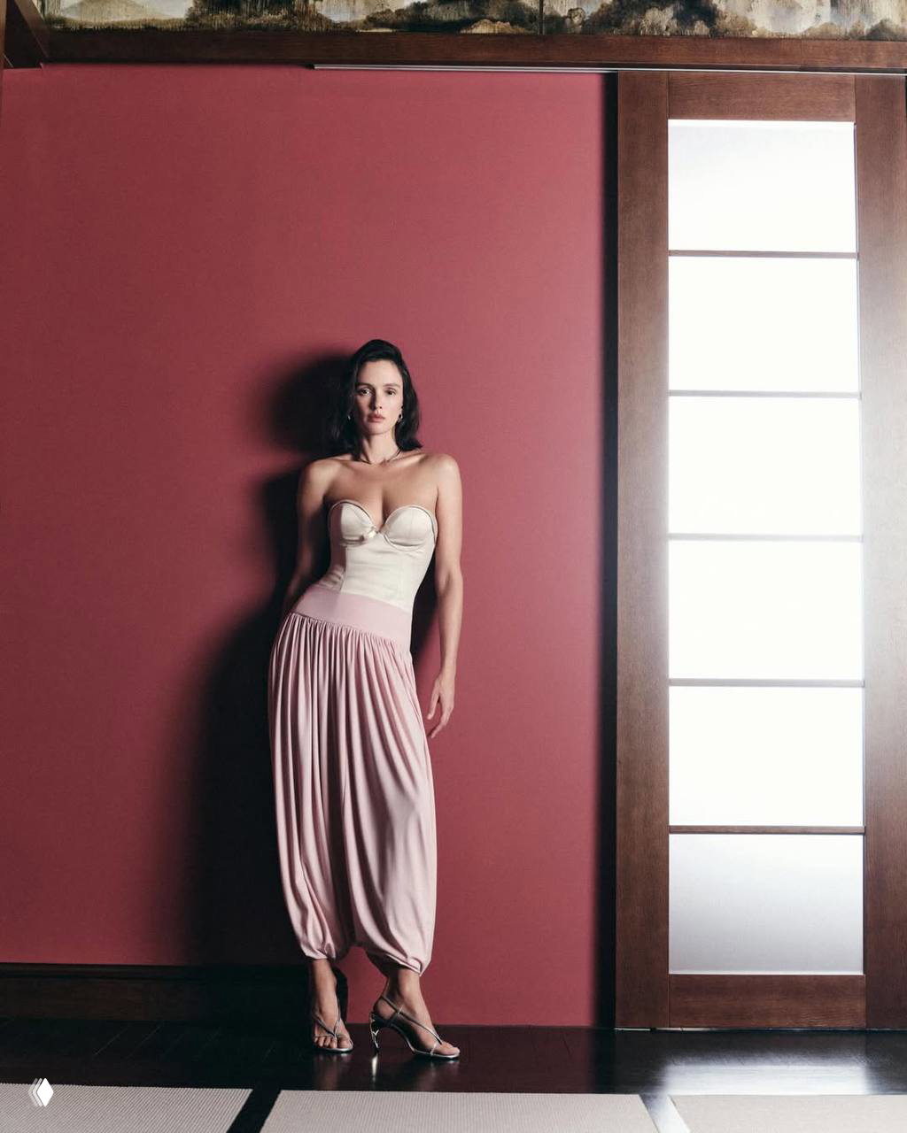 Паулина Андреева в полный рост у бордовой стены в светлом корсете и розовых драпированных брюках, образ в пастельной гамме.