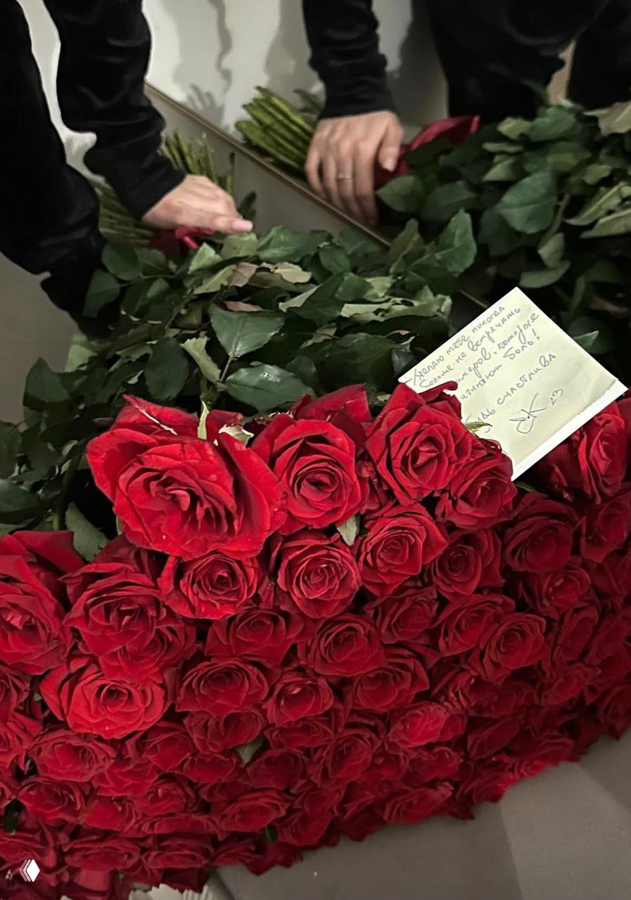 Крупный план большого букета красных роз с бумажной запиской, на которой заметна подпись «EK»; листья и стебли заполняют кадр.