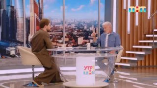 Телевизионная студия: двое мужчин за стеклянным столом на фоне городской панорамы и декора, интервью в утренней передаче.