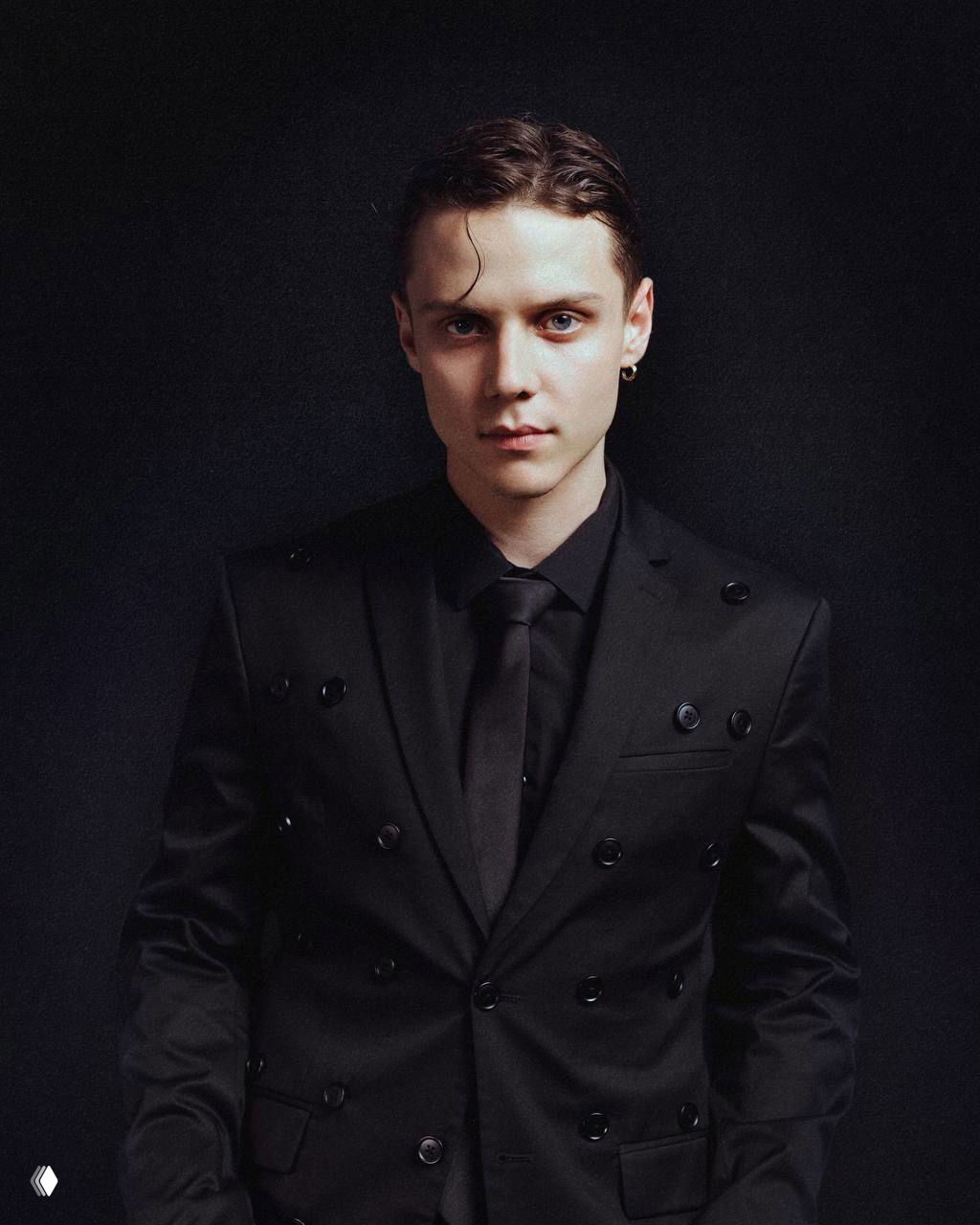 Формальный студийный портрет в тёмном костюме и галстуке — строгий образ артиста в классическом стиле с драматическим светом.