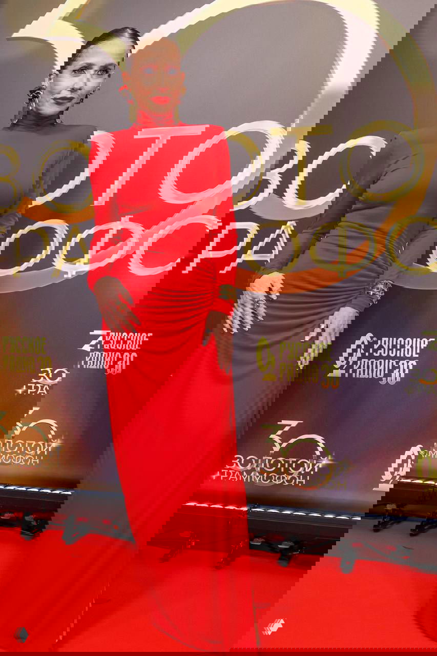 Юлия Барановская в длинном красном платье с высоким воротником и характерным макияжем стоит на красной дорожке церемонии «Золотой Граммофон» — фото с премии 30 лет Русского Радио.