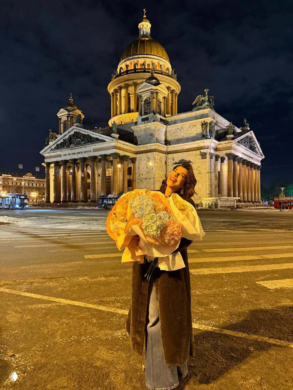 Екатерина Волкова на ночной улице с большим букетом цветов на фоне подсвеченного собора; уличный городской пейзаж и праздничное настроение видны в кадре.