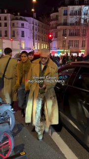 Ночной уличный кадр в Париже: пара в тёплой одежде идёт по тротуару возле припаркованных машин и светофора после показа.