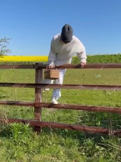 Кадр из видео: человек в светлой одежде перелезает через деревянный забор с корзиной, виден зелёный луг и жёлтые цветы на горизонте.
