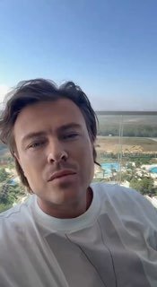 Селфи мужчины на балконе отеля с видом на бассейн и побережье; вертикальное фото при дневном свете, нейтральное выражение лица, городской пейзаж за спиной.