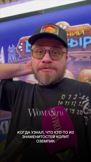 Крупный план Гарика Харламова в кепке и очках, он держит микрофон Woman.ru на фоне яркого баннера, говорит в камеру.