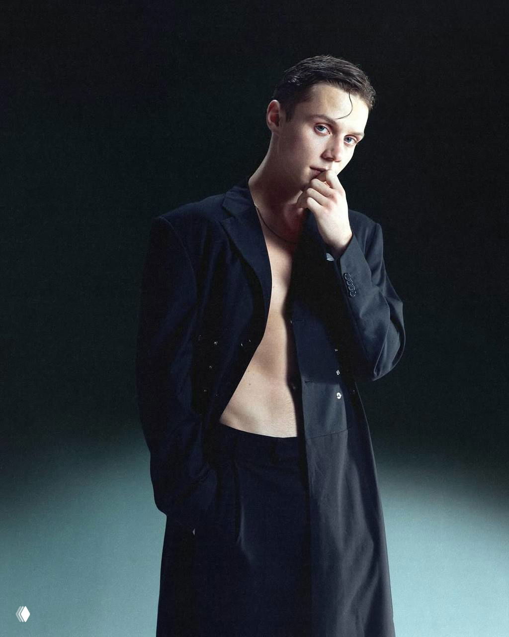 Полуобнажённый студийный портрет в пиджаке на тёмном фоне: артист стоит в задумчивой позе, подчеркнутый контрастами света и тени.