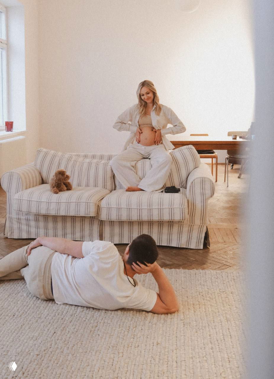 Даша сидит на диване, держит живот, вокруг светлая минималистичная комната; Никита лежит на полу и смотрит на неё — домашняя сцена ожидания.