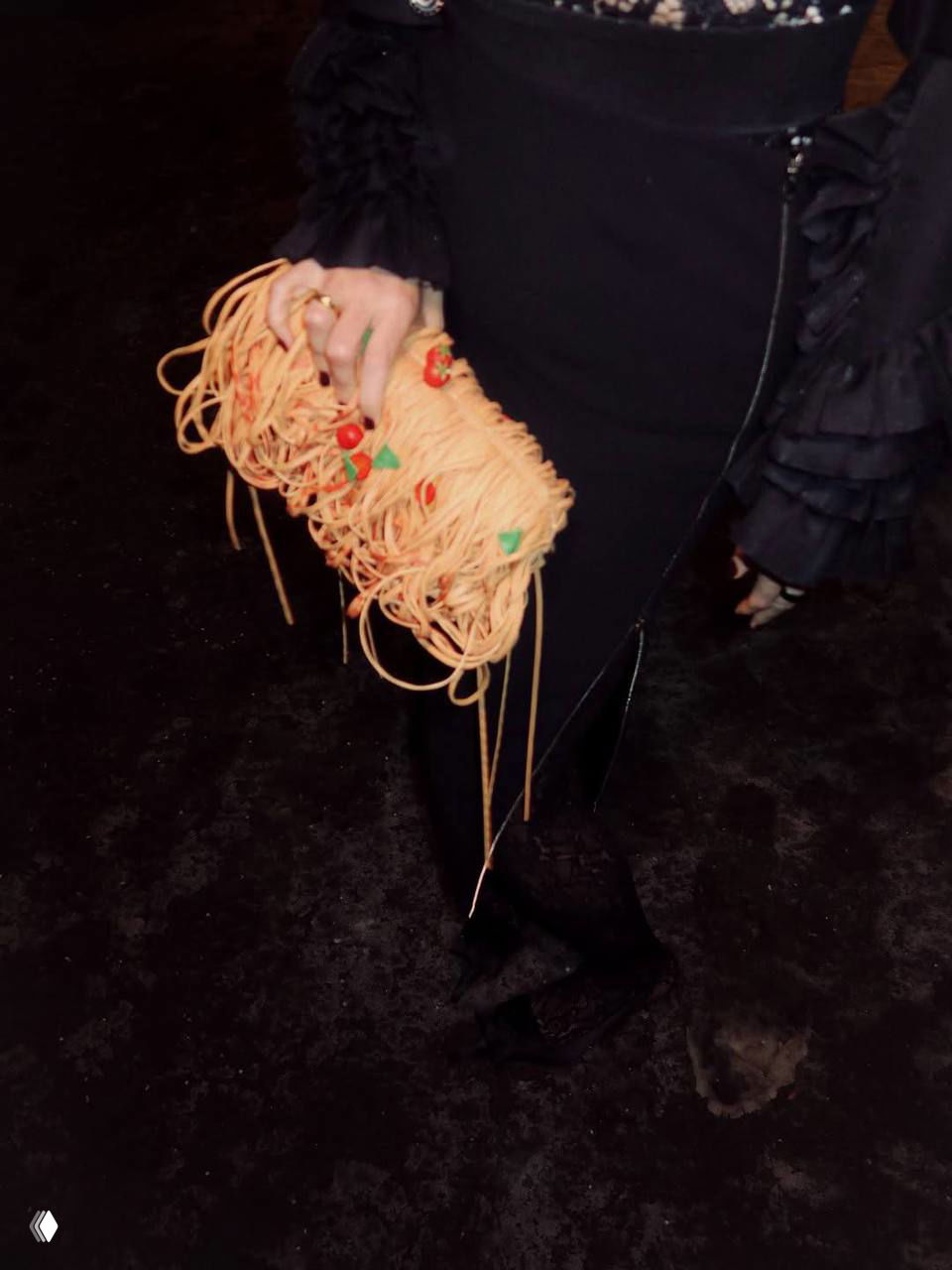 Крупный план рук в платье, держащих сумку‑«лапшу» с декоративными элементами и свисающими нитями, видны детали аксессуара.