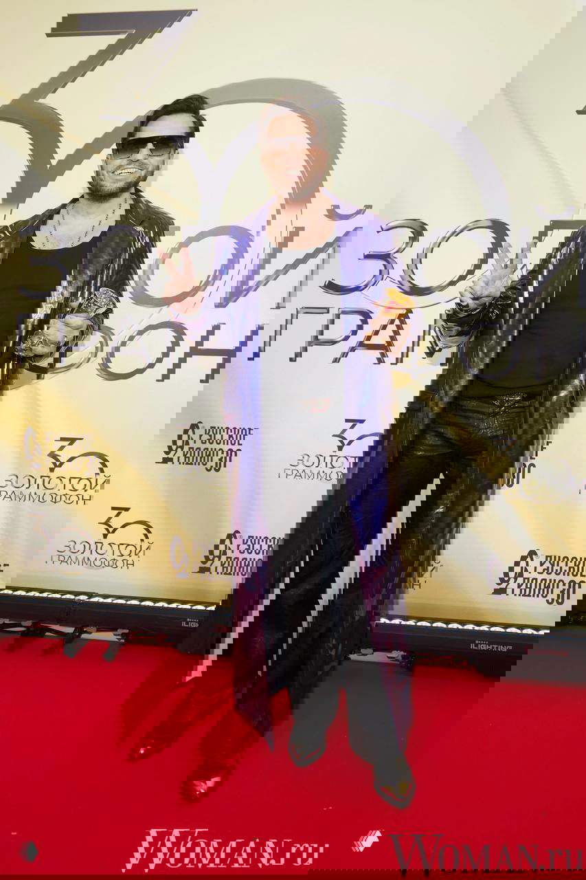 Александр Ревва в блестящем фиолетовом плаще и тёмной футболке позирует с трофеем на красной дорожке на фоне брендирования мероприятия.