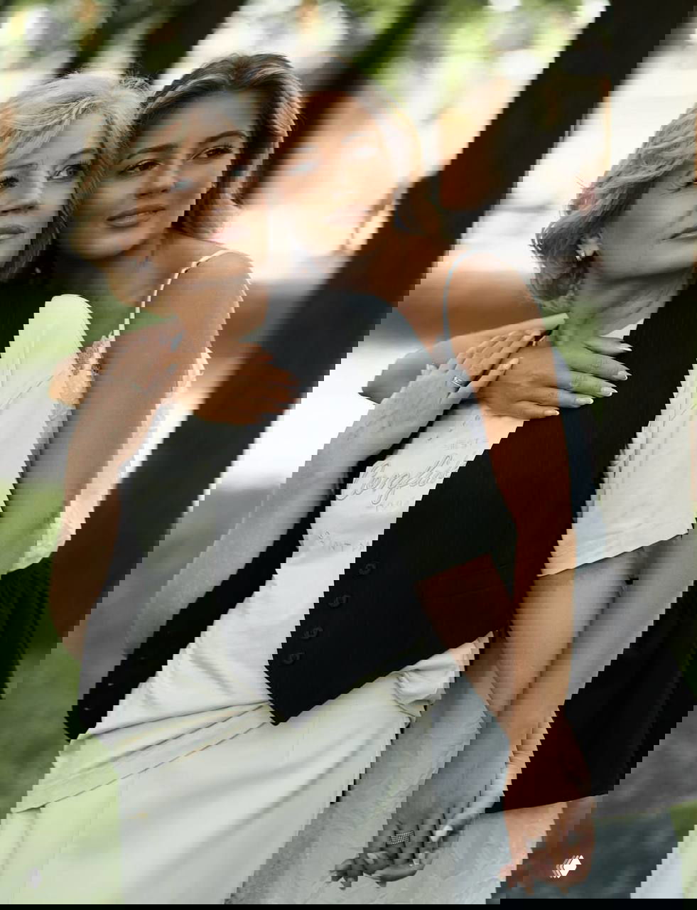 Молодая женщина обнимает пожилую даму на открытом воздухе: семейный кадр на зелёном фоне, выражение нежности и поддержки между поколениями.