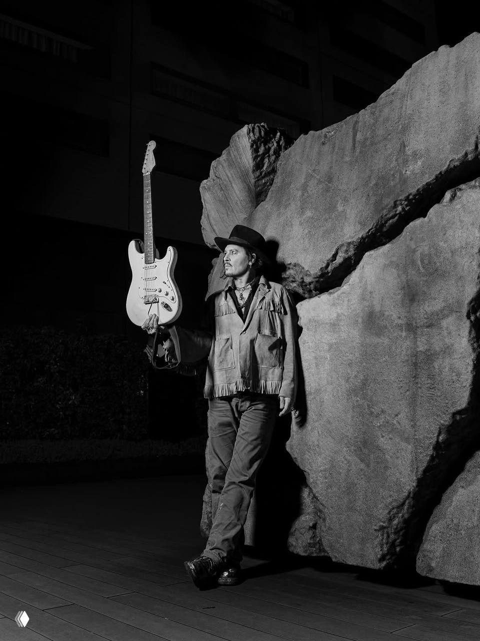 Художественный чёрно-белый портрет в Токио: мужчина в шляпе опирается о камень и держит электрогитару, городской ночной фон и мягкие тени создают винтажный образ