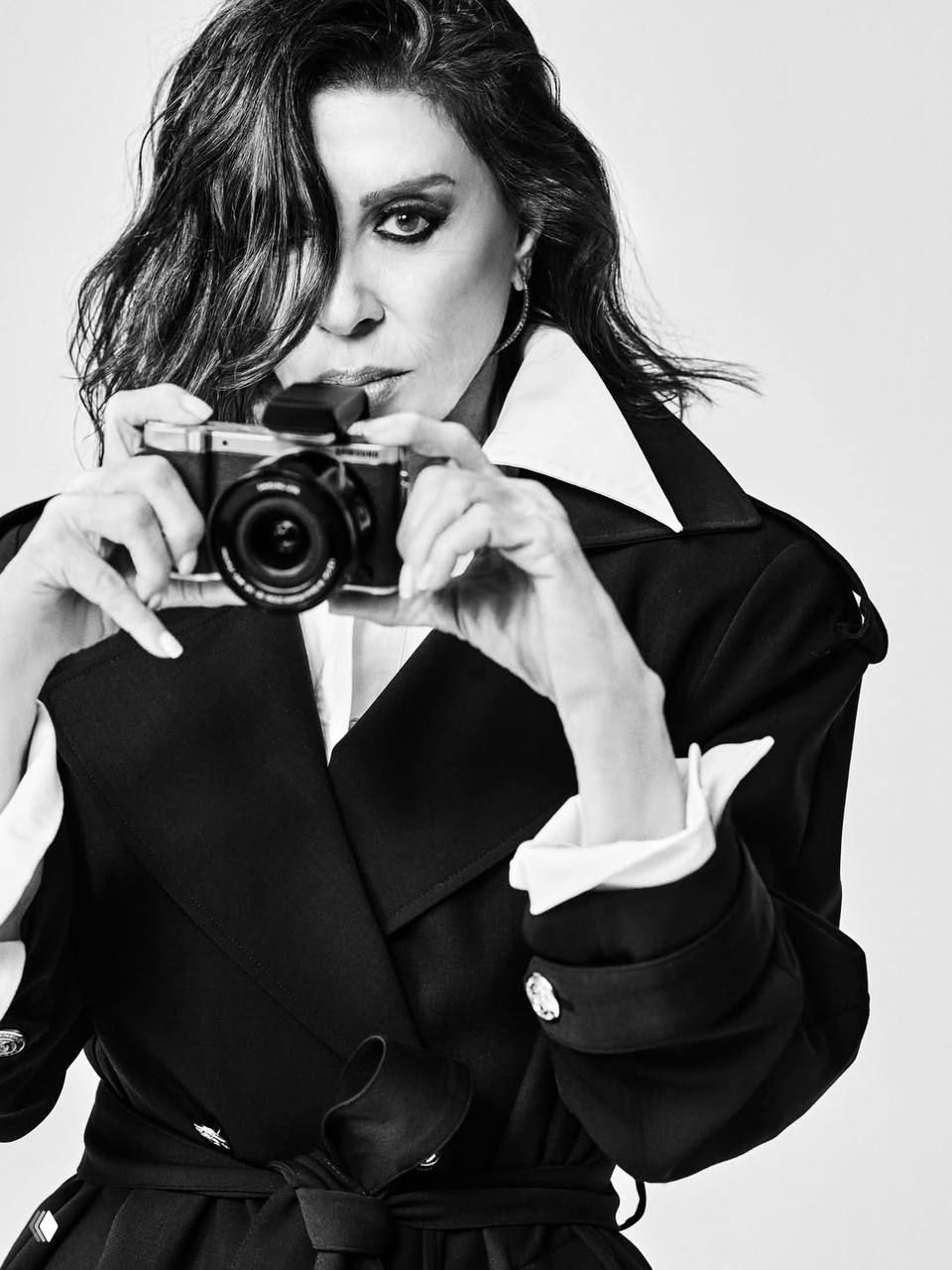 Крупный чёрно‑белый план: женщина держит фотоаппарат близко к лицу, выразительный макияж и прямой, сильный взгляд