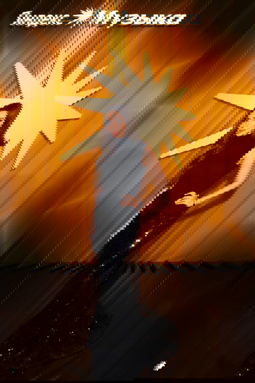 Длинноплатьная темноволосая женщина в элегантном образе стоит у золотой инсталляции с надписью «Яндекс Музыка», фотографы вокруг.