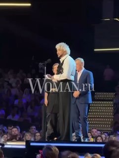 Филипп Киркоров на сцене во время выступления у микрофона, видна сцена, зрительный зал и водяной знак Woman.ru — кадр с награждения/концерта.