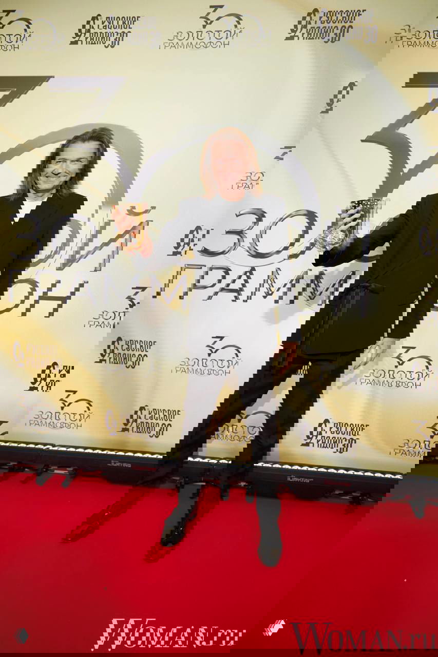 Дмитрий Маликов в чёрном пиджаке с поясом улыбается, демонстрируя награду на красной дорожке юбилея «Русского радио», фотографы вокруг.