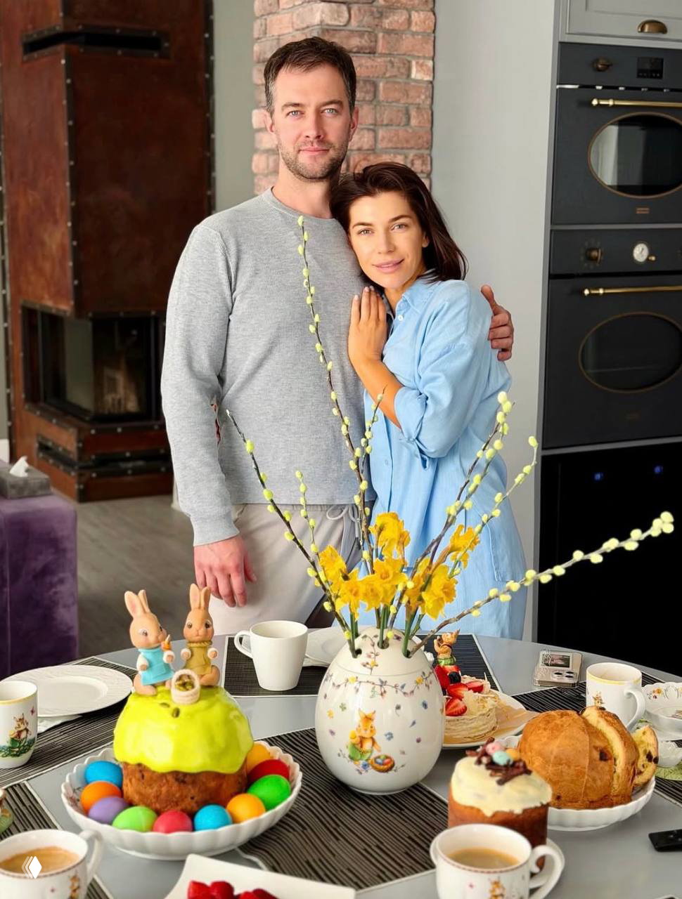 Пара обнимается на фоне пасхального стола: на столе куличи, чашки и праздничные детали, домашняя атмосфера и весенний декор.