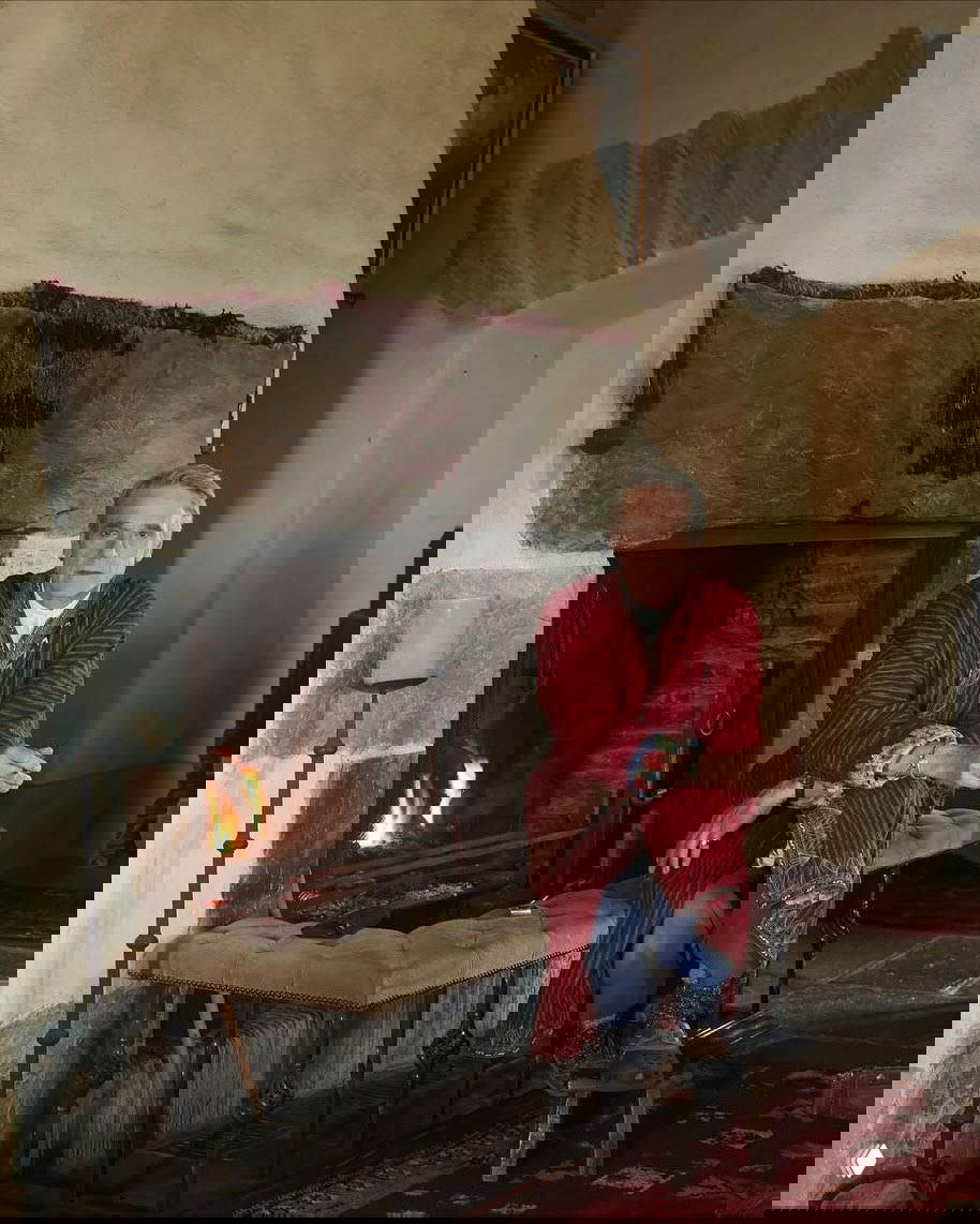 Джереми Айронс сидит у камина в расслабленной позе в традиционной одежде, рядом горит огонь и видны элементы старинного очага замка.