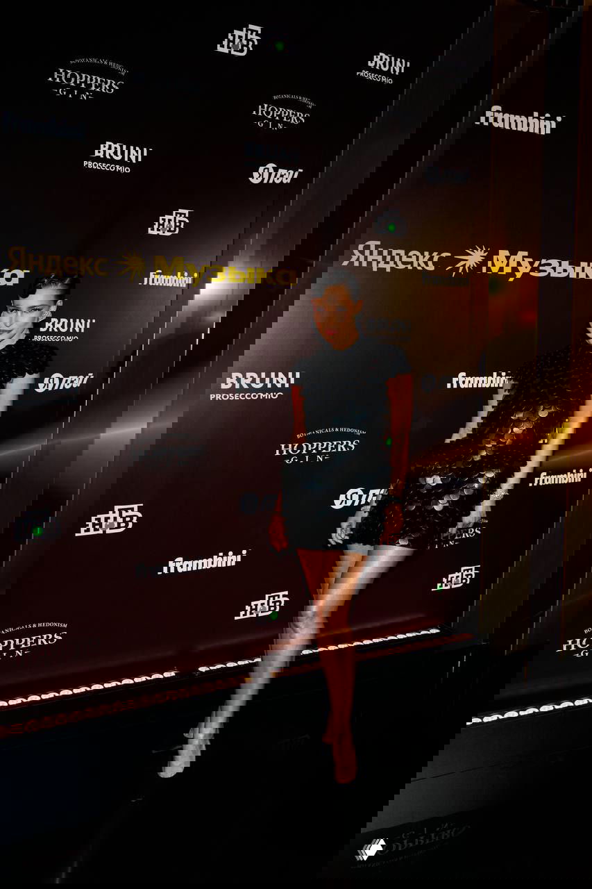 Темноволосая женщина в коротком черном платье с текстурой стоит на красной дорожке премии на фоне брендированных панелей «Яндекс Музыка».