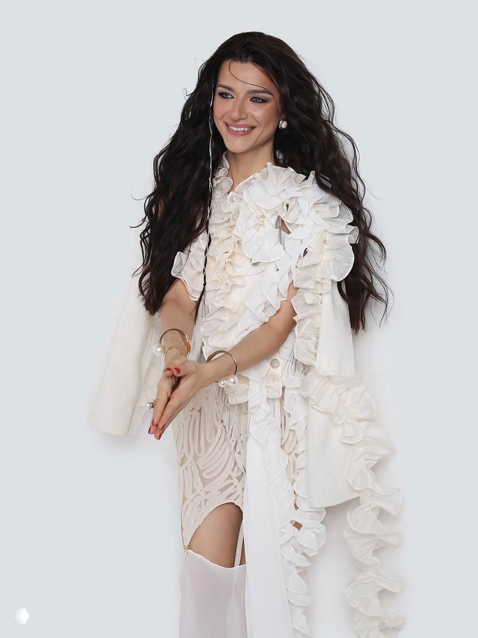 Женщина в белом платье с рюшами в полный рост на светлом фоне, поза для фотосессии, видны украшения и текстура наряда