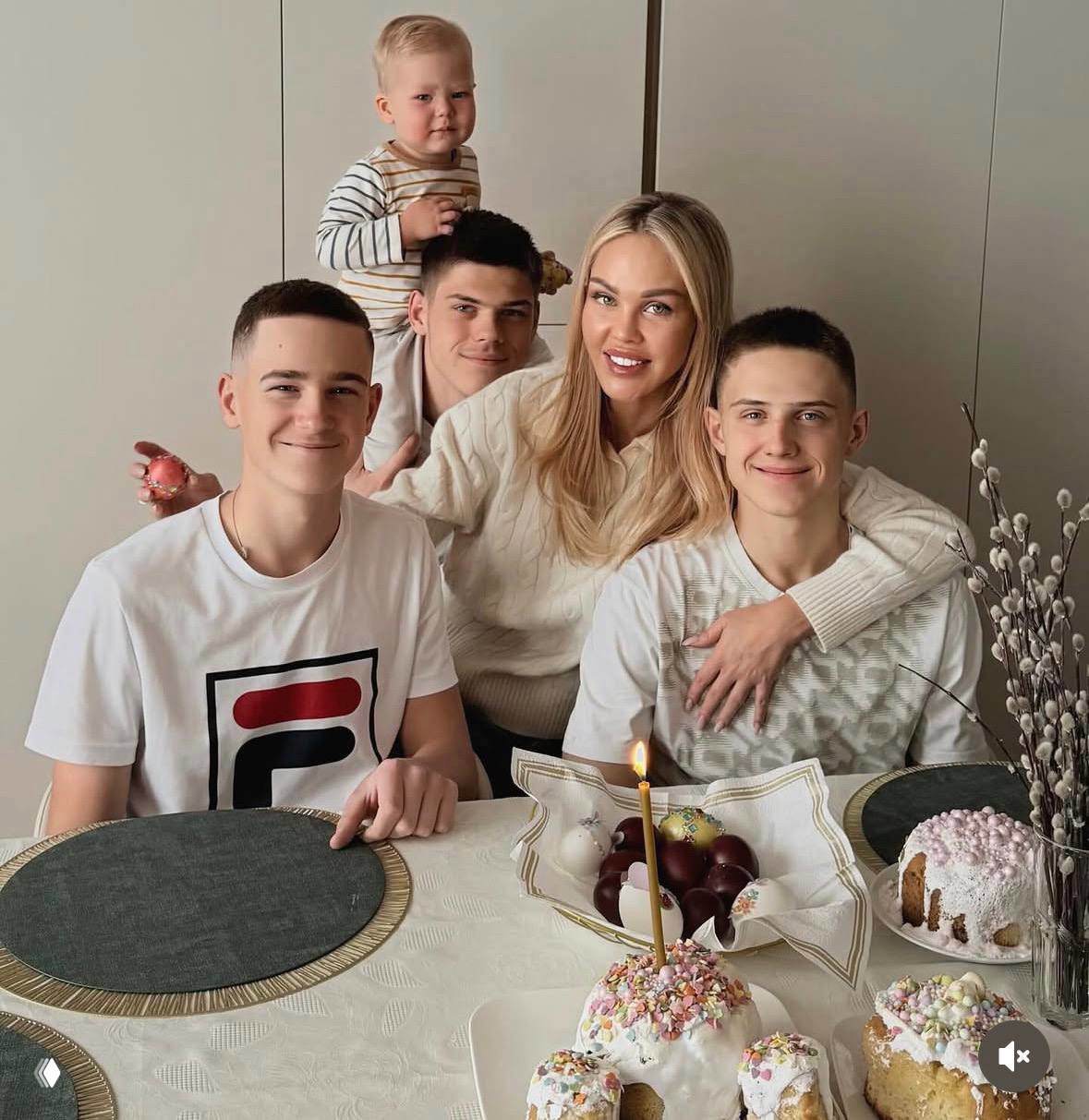 Семейный снимок за столом: женщина с тремя подростками и малышом на плечах, на столе пасхальные десерты, свеча и праздничная сервировка.