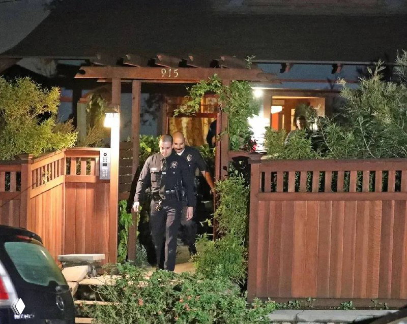 Ночное уличное фото: два сотрудника полиции в форме выходят через деревянную калитку у дома с подсветкой и растительностью у забора.