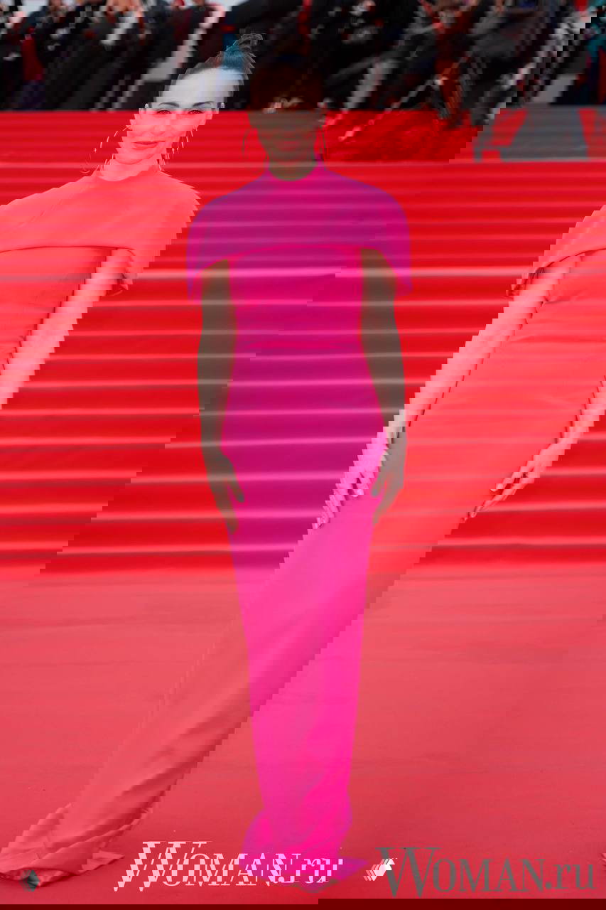 Женщина в ярком розовом платье-футляре позирует на красной дорожке 48-го ММКФ на фоне лестницы и прессы, строгая укладка волос.