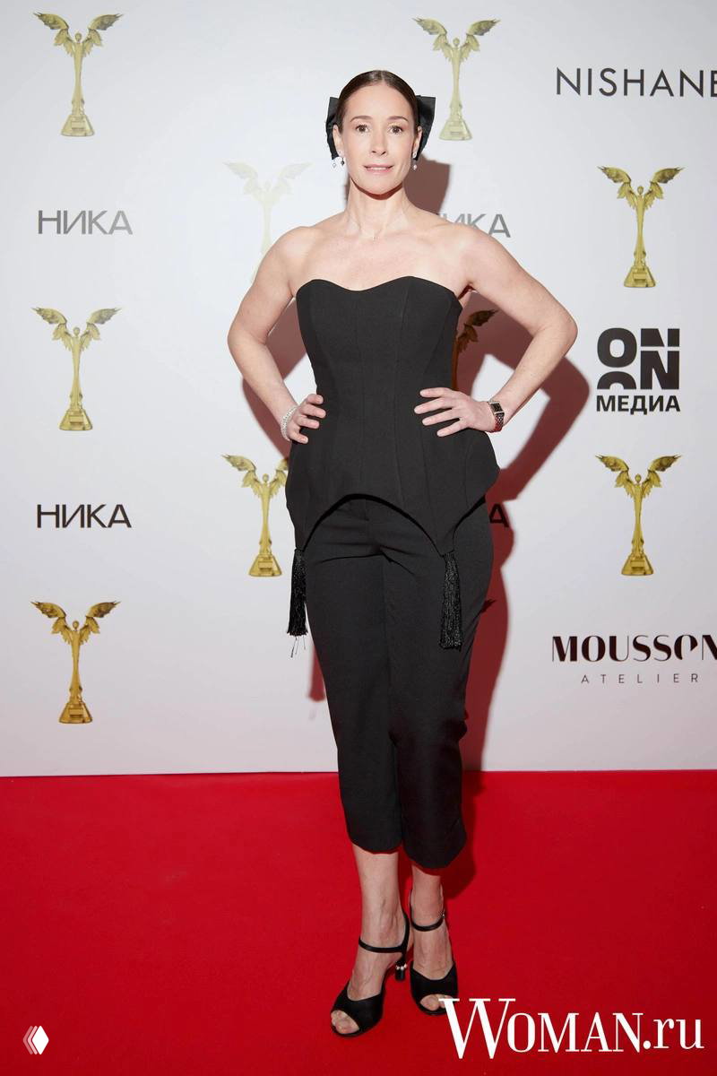 Женщина в чёрном корсетном костюме и укороченных брюках на красной дорожке премии «Ника», на фоне логотипов.