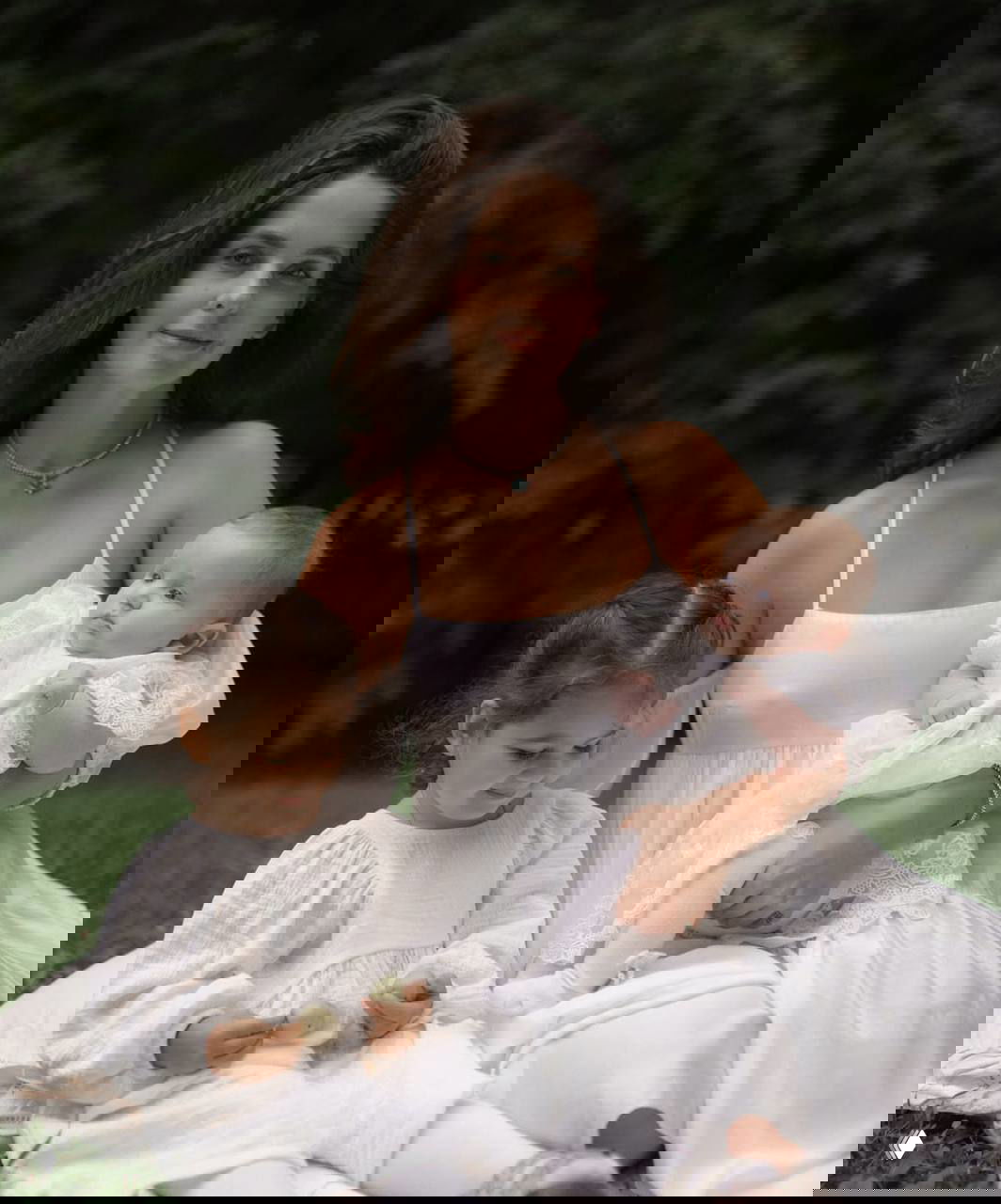 Молодая женщина сидит на траве с младенцем и ещё одним ребёнком: нежный семейный портрет на природе в светлых платьях, спокойная сцена.