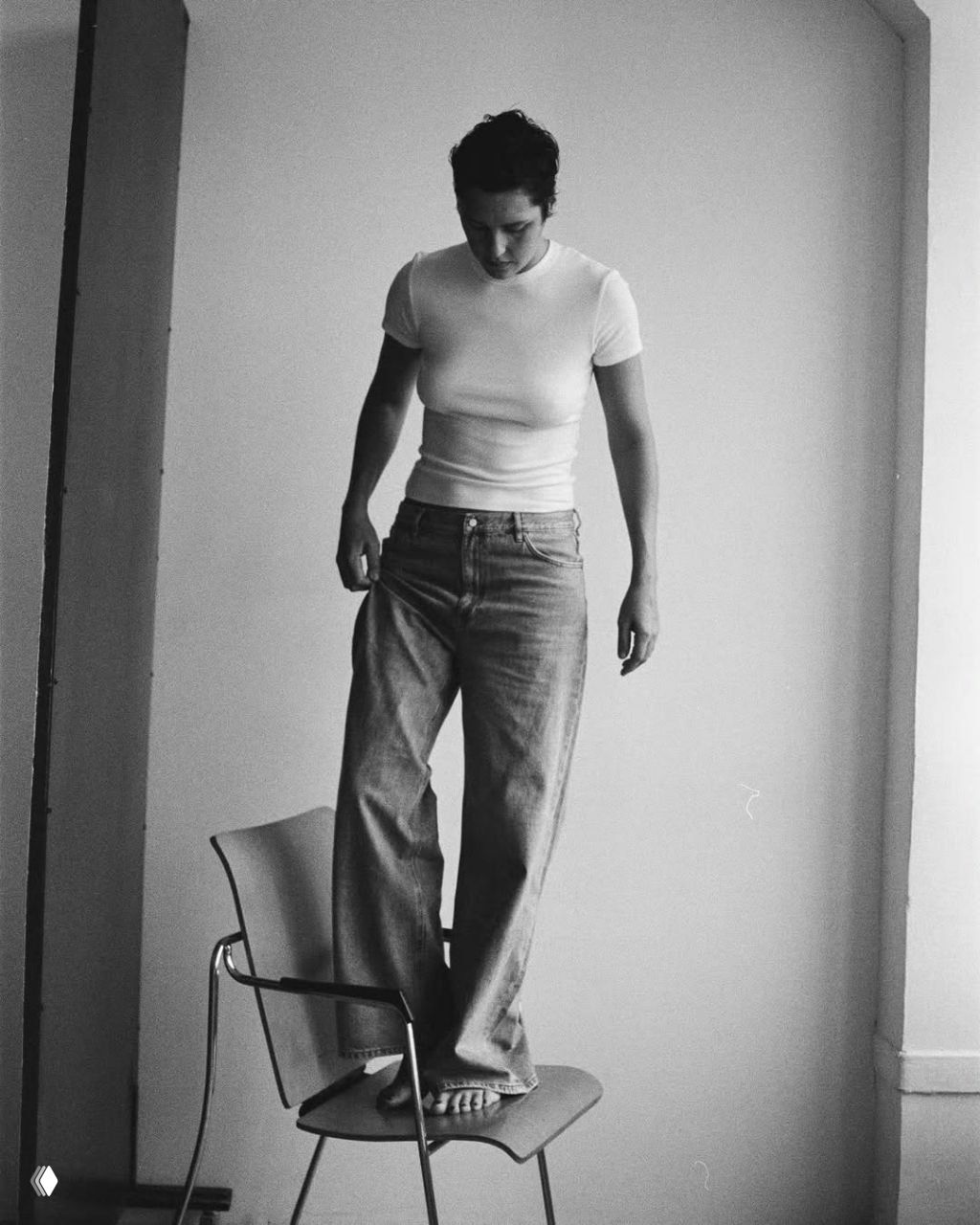Чёрно‑белый кадр: женщина стоит на стуле в джинсах и футболке, минималистичная студия и вертикальная композиция.