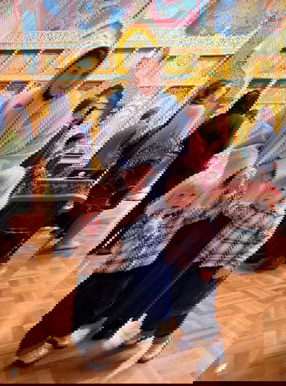 Посещение храма: женщина в платке стоит с двумя маленькими детьми на фоне иконостаса, прихожане и внутреннее убранство церкви видны вокруг.