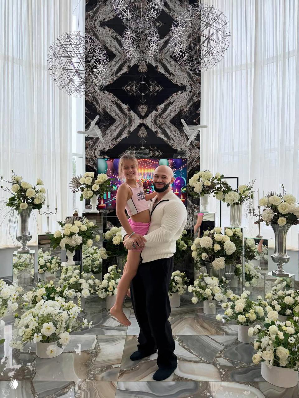 Джиган держит на руках дочь Майю на фоне гостиной, усыпанной белыми цветочными композициями и большим декоративным панно за ними.