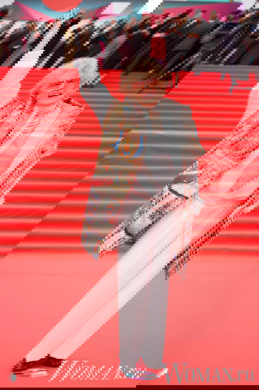 Пожилая женщина в светлом костюме и украшениях радостно машет рукой на красной дорожке 48-го ММКФ у входа в театр «Россия», вокруг фотографы.