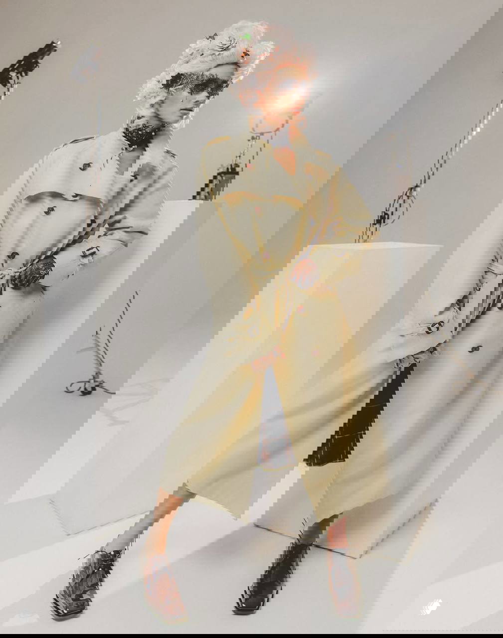 Модель в кремовом тренче и розовом парике с рококо-аксессуарами, позирует у белых подиумов: видна студийная аппаратура и световые приборы.