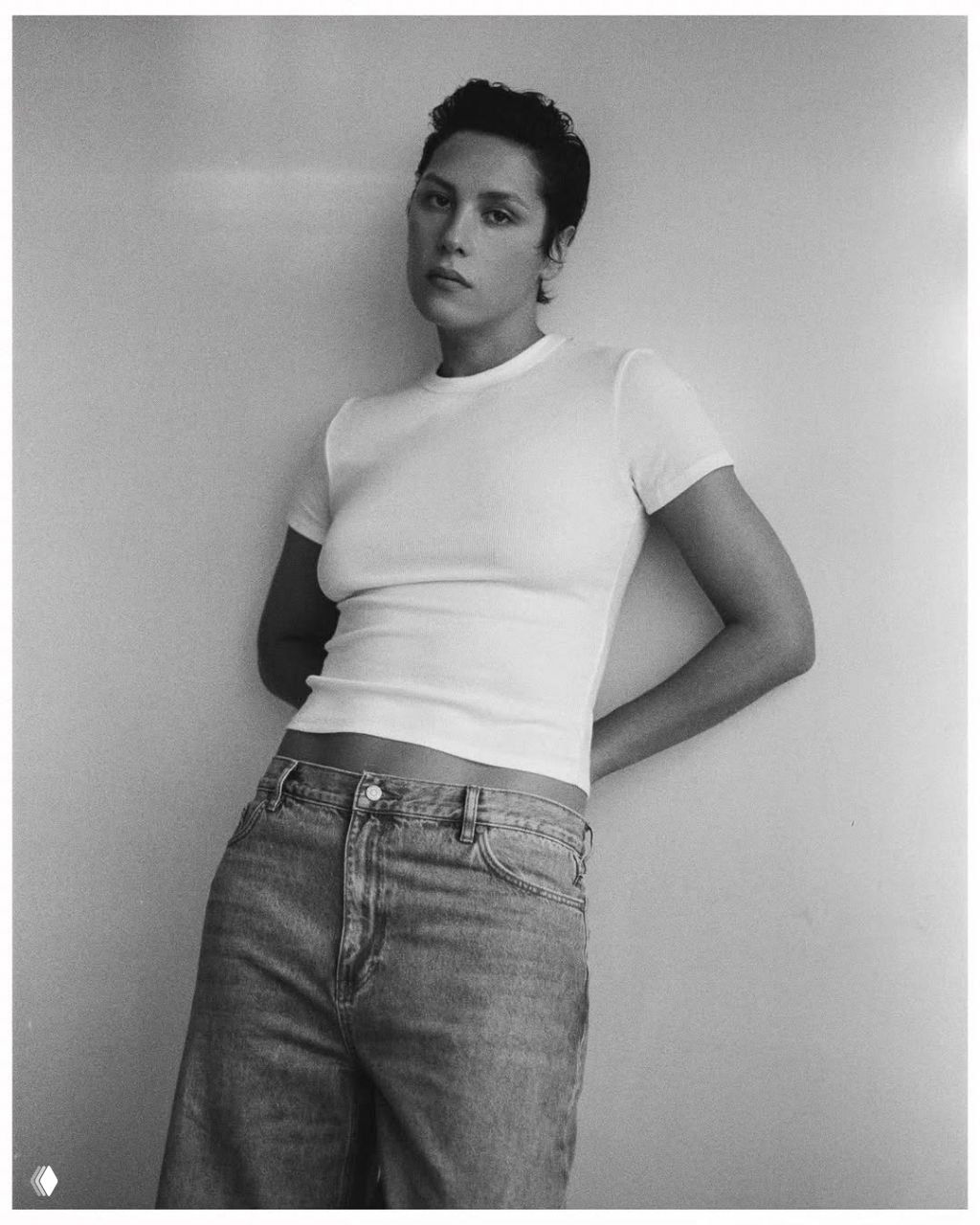 Чёрно‑белый портрет в полный рост: женщина в футболке и джинсах, опирается о стену на нейтральном фоне студии.